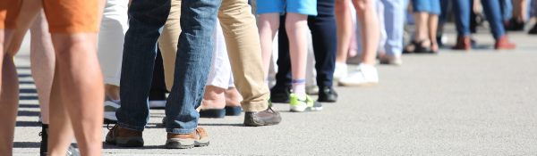 A photo taken from a low angle, showing the knees and below of people in a long line waiting to get into an event.